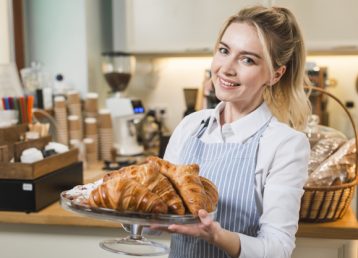 panadero-femenino-sonriente-que-sostiene-croissant-cocido-soporte-torta_23-2148027951